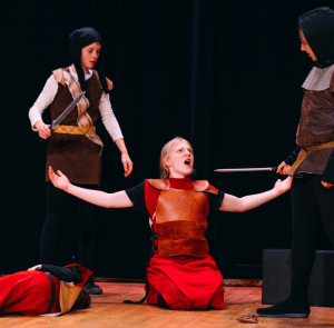 A kneeling actor is surrounded by a soldier on either side, each holding long swords. The kneeling actor is surrendering bravely.