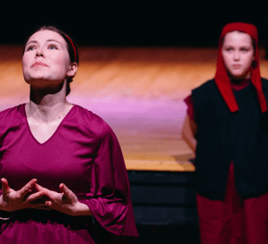 A young actor expressing a deep emotion, looking up at the sky, while her servant looks after her in the background.